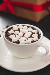 Fototapeta na wymiar Hot chocolate drink in white cup with marshmallow and christmas gift box on dark background