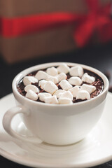Hot chocolate drink in white cup with marshmallow and christmas gift box on dark background