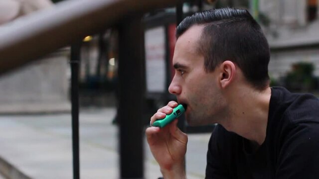 Close up of a guy smoking a green vape outside on the steps
