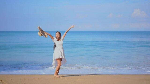 A pretty young woman walking along a sandy beach toward the sea reacts in joy by twirling around in circles.