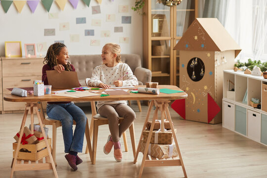 Full Length Portrait Of Two Smiling Girls Crafting Together While Sitting In Decorated Playroom, Copy Space