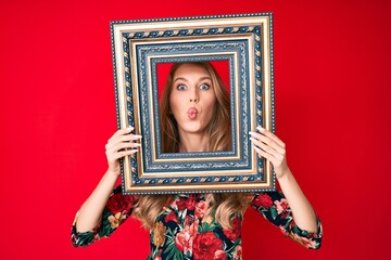 Fototapeta premium Young caucasian woman with blond hair holding empty frame puffing cheeks with funny face. mouth inflated with air, catching air.