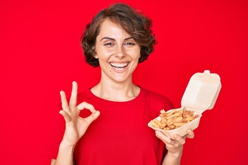 Young hispanic woman holding potato chip doing ok sign with fingers, smiling friendly gesturing excellent symbol