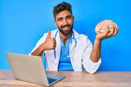 Young hispanic man wearing doctor uniform working at the clinic holding brain smiling happy and positive, thumb up doing excellent and approval sign
