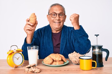 Fototapeta na wymiar Senior handsome man with gray hair sitting on the table eating croissant for breakfast screaming proud, celebrating victory and success very excited with raised arms