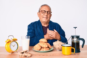 Senior handsome man with gray hair sitting on the table eating breakfast in the morning smiling with hands on chest, eyes closed with grateful gesture on face. health concept.