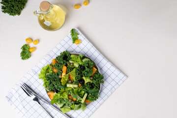 veggie salad of lettuce leaves and kale. Yellow tomatoes, oruganets, olives and capers. A plate on a checkered cloth napkin. A bottle of olive oil is nearby. Light vegetable salad. Light background.