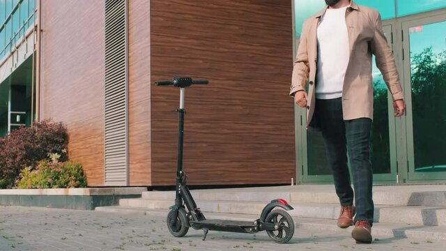 Full shot of handsome successful businessman in smart clothes finished talking on phone and leaving modern glass office on electric scooter in daylight