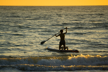 Young guy in the sea at sunset, mastering the technique of surfing, stands up with a paddle