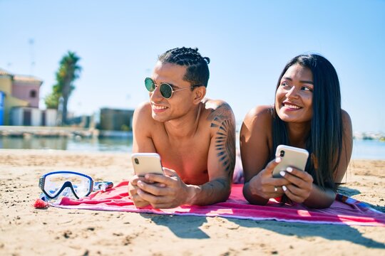 Young latin couple using smartphone lying on the sand at the beach.