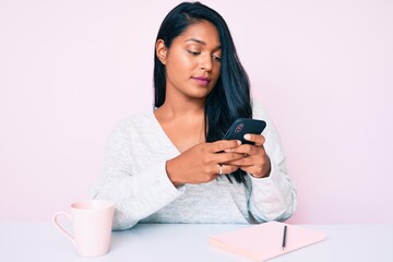 Fototapeta premium Beautiful latin young woman with long hair using smartphone sitting on the table thinking attitude and sober expression looking self confident