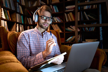 Young business man, male student wearing headphones elearning on laptop computer sitting in chair working from home office, learning online, studying remote training course, watching webinar stream.