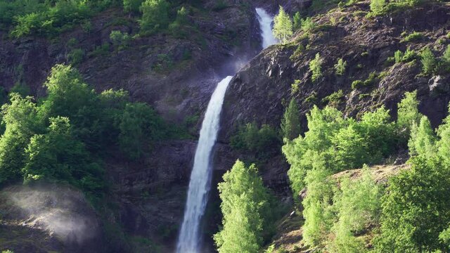 One of many waterfalls in the Naeroy fjord shores. A powerful torrent of water falling from the cliff, raising foam and water spray in the air. Lush green vegetation covers the mountainsides.