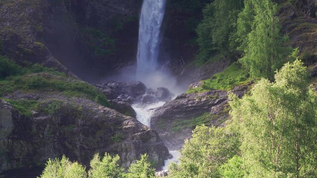 One of the waterfalls in the Naeroy fjord shores. A powerful torrent of water falling from the cliff, raising foam and water spray in the air. Lush green vegetation surrounds the waterfall.