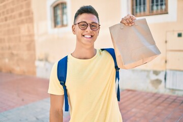 Fototapeta premium Young hispanic student smiling happy holding take away paper bag at street of city