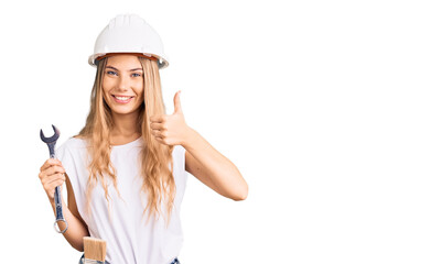 Beautiful caucasian woman with blonde hair wearing hardhat and builder clothes smiling happy and positive, thumb up doing excellent and approval sign