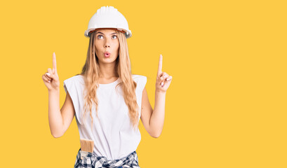Fototapeta na wymiar Beautiful caucasian woman with blonde hair wearing hardhat and painter clothes amazed and surprised looking up and pointing with fingers and raised arms.