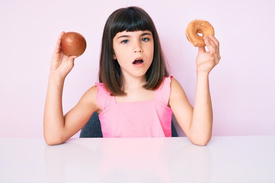 Young little girl with bang holding red apple and donut sitting on the table in shock face, looking skeptical and sarcastic, surprised with open mouth