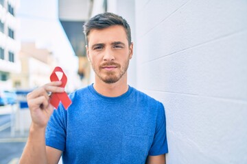 Young caucasian man with serious expression holding red hiv ribbon at the city. - obrazy, fototapety, plakaty