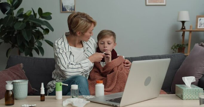 Young woman talking and giving thermometer to her teen kid, showing pills during video conversation. Mom and son looking at laptop screen while having online consultation with doctor
