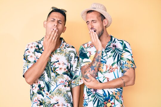 Young gay couple of two men wearing summer hat and hawaiian shirt bored yawning tired covering mouth with hand. restless and sleepiness.