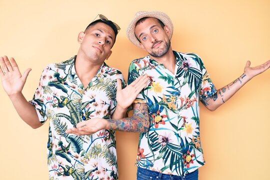 Young gay couple of two men wearing summer hat and hawaiian shirt clueless and confused expression with arms and hands raised. doubt concept.