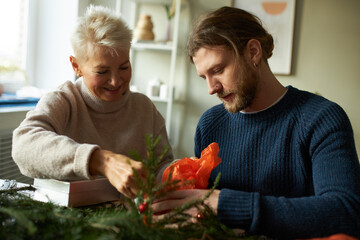 Cozy scene of two generations bonding while preparing for winter holidays. Joyful senior female and young unshaven man in warm sweaters sitting at table with spruce twigs and boxes, making presents