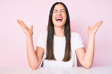 Young caucasian woman sitting at the table wearing casual white tshirt celebrating mad and crazy...