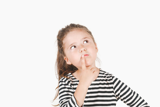 Thoughtful girl looking up aside and touching her mouth with finger. Girl with eyes directed sideways and head tilted to one side. Posing little girl wearing striped shirt. Isolated background.