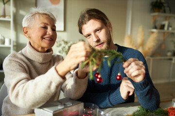 Portrait of stylish young bearded man helping his senior mother decorating house. Attractive happy retired lady and her unshaven son hanging shiny New Year ornaments on green coniferous branch