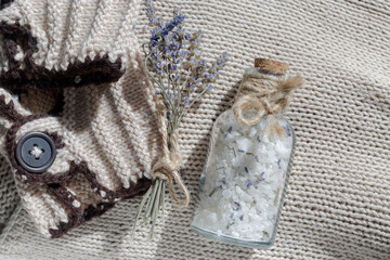 A glass bottle of sea salt with a cork for a relaxing bath on a beige textile knitted background next to a small bouquet of lavender and a body scrub