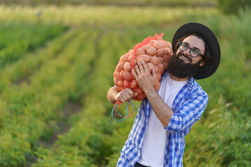 Smiling young farmer holding a sack of fresh potatoes on green potato field background.