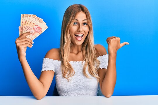 Beautiful blonde young woman holding 100 mexican pesos banknotes pointing thumb up to the side smiling happy with open mouth