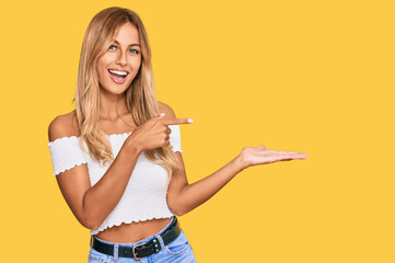 Beautiful blonde young woman wearing casual white tshirt amazed and smiling to the camera while presenting with hand and pointing with finger.