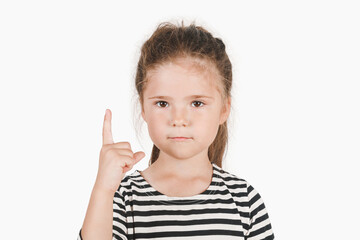 Serious girl pointing finger up, showing Number one. Close up of posing little girl wearing a striped shirt, looking at the camera on an isolated background. First, the winner, attention concept.