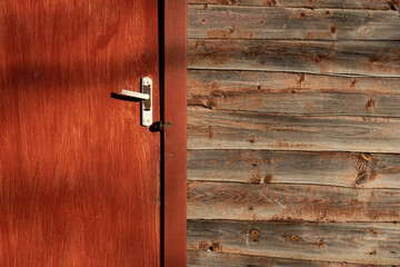 Fragment of the wall of a rustic wooden house. There is a brown door with a white rusted handle and...