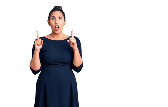 Young beautiful brunette woman wearing casual dress amazed and surprised looking up and pointing with fingers and raised arms.