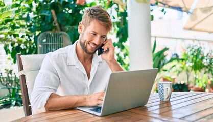 Handsome caucasian man sitting on the terrace working from home using computer laptop and speaking on the phone