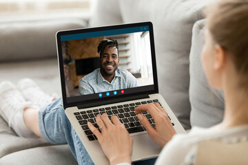 Happy young european woman relaxing on sofa, holding web camera video call with smiling handsome african american boyfriend, using computer software application, distant communication concept.