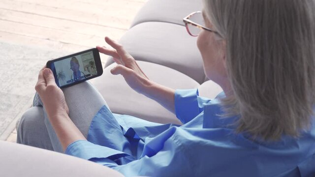 Over shoulder view of older elderly woman patient meeting african virtual doctor using mobile phone at home. Online telemedicine visit. Seniors ehealth, telehealth medical video call consultation.