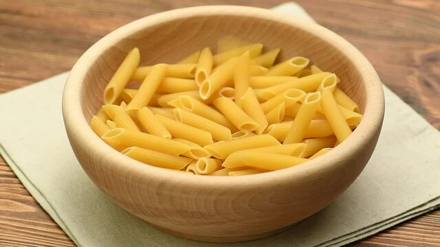 Dry raw pasta penne rigate falling into a wooden bowl, slow motion