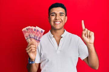 Young arab man holding south african rand banknotes smiling with an idea or question pointing finger with happy face, number one