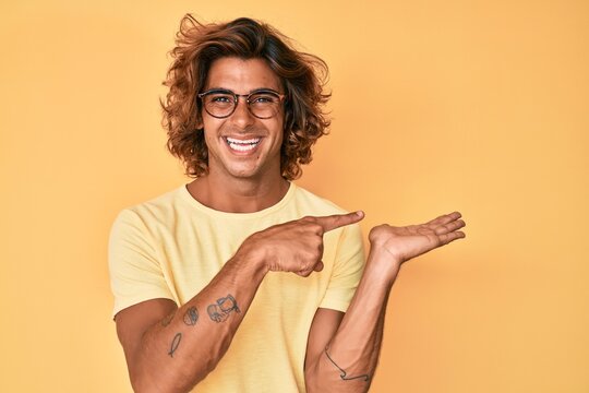 Young hispanic man wearing casual clothes and glasses amazed and smiling to the camera while presenting with hand and pointing with finger.