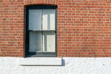 A vintage exterior red brick wall with a black wooden double hung window to the left of the wall. The ledge of the window is white and the brick wall below the window is painted with white paint. 