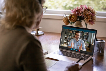 Back rear view middle aged older woman holding online conversation with grown up son or enjoying distant educational class with young male tutor using video call computer application at home.