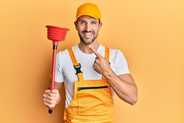Young handsome man wearing plumber uniform holding toilet plunger smiling happy pointing with hand and finger