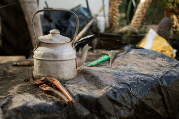 an old, dusty aluminum teapot on a dirty tarp. Next to it are pruning shears, a screwdriver, and a paper knife. Photo taken in the village