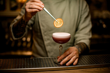 bartender holds tweezers with slice of orange over wine glass with foam cocktail