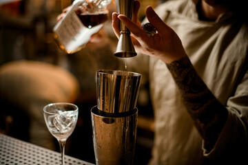Close-up view on barman pours drink from metal jigger into shaker on bar counter