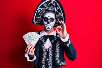 Young man wearing mexican day of the dead costume holding dollars doing ok sign with fingers, smiling friendly gesturing excellent symbol